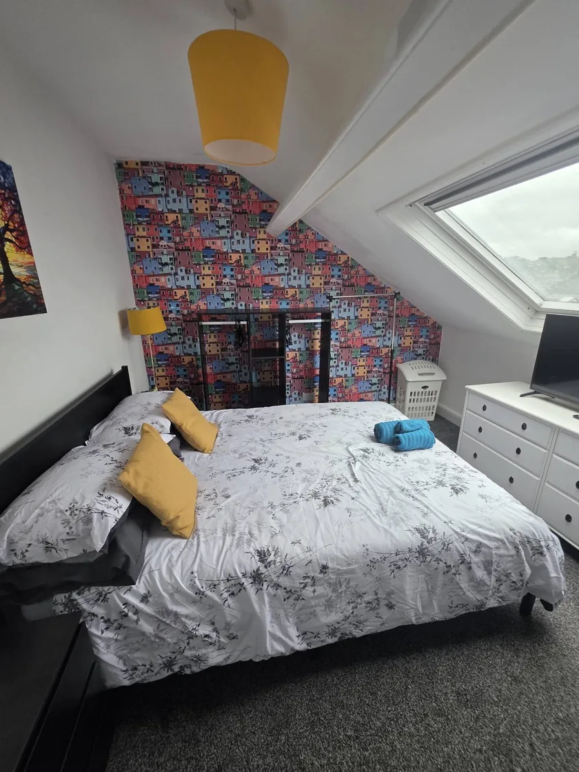 Bed beneath a sloping ceiling, with yellow cushions and colourful house-patterned wallpaper.