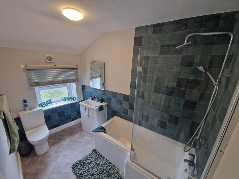 Bathroom with a bath, overhead and handheld shower, glass screen, basin and blue-green wall tiles.
