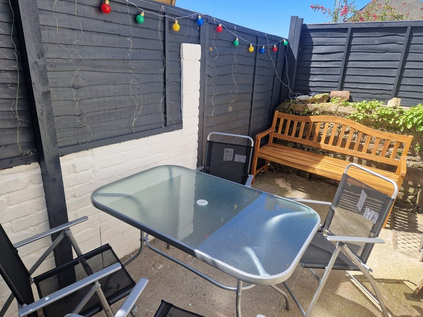 Paved courtyard with a glass-topped table, outdoor chairs, wooden bench and coloured string lights.