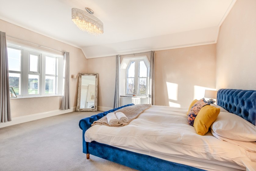 A blue upholstered bed with white linen and a yellow cushion in a pale room with two windows and a leaning mirror.