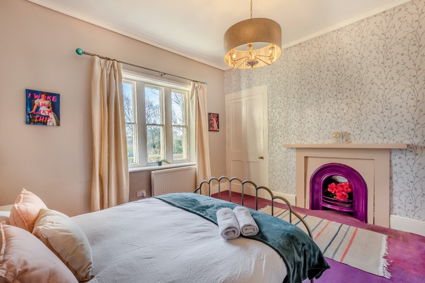 A metal-framed bed with white linen and a teal throw in a room with a purple fireplace, patterned wallpaper and a large window.