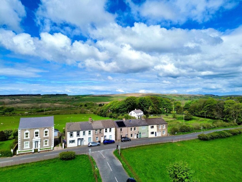 Aerial view of a short row of cottages beside open green fields and a country road.