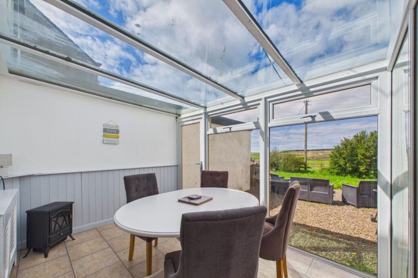 Round dining table and upholstered chairs beneath a glazed conservatory roof, opening towards the garden.