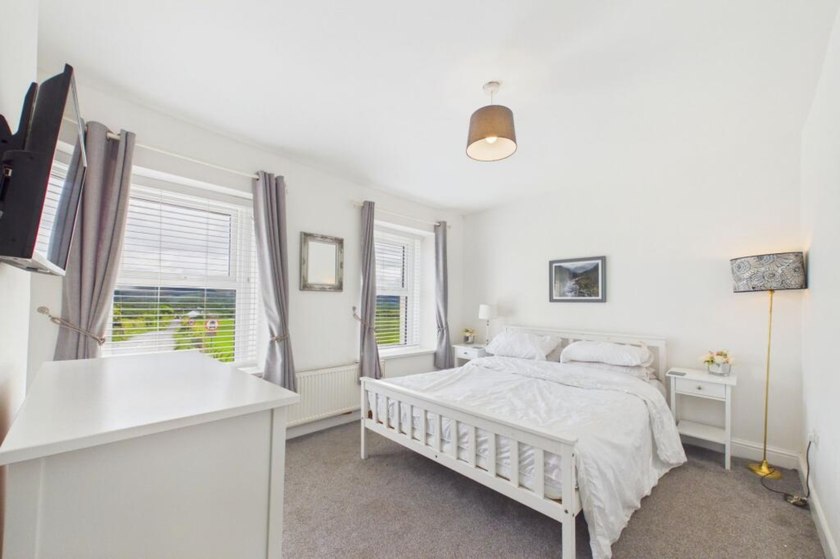 White double bed in a light bedroom with a dresser, bedside tables and countryside views through the windows.