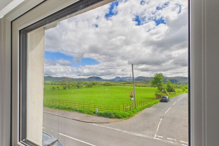 View through a cottage window across a country road towards green fields and distant hills.